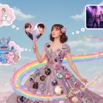 A young woman with brown hair, wearing a shimmering pink dress adorned with numerous fandom-related buttons and trinkets, stands on a cloud with a blue sky above